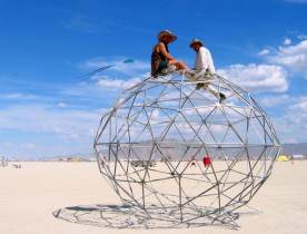 Men climb geodesic globe with kite festival in background
